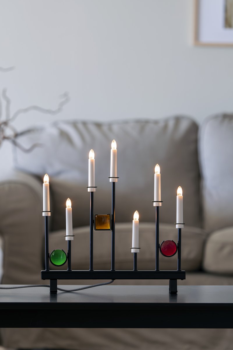 Elljusstake 7ljus Svart från Konstsmide - De sju ljusen ramas in av dekorativa f&auml;rgade glasdetaljer i gr&ouml;nt, r&ouml;tt och b&auml;rnstensgult, vilket skapar en elegant och festlig atmosf&auml;r.