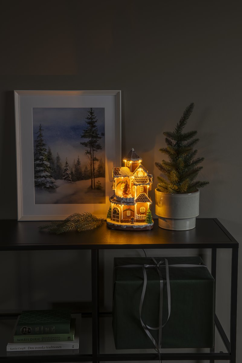Pepparkaksby 30cm Brun från Konstsmide - Det detaljrika huset med sn&ouml;t&auml;ckta tak, lysande f&ouml;nster och dekorerad gran sprider ett varmt och inbjudande sken i rummet.