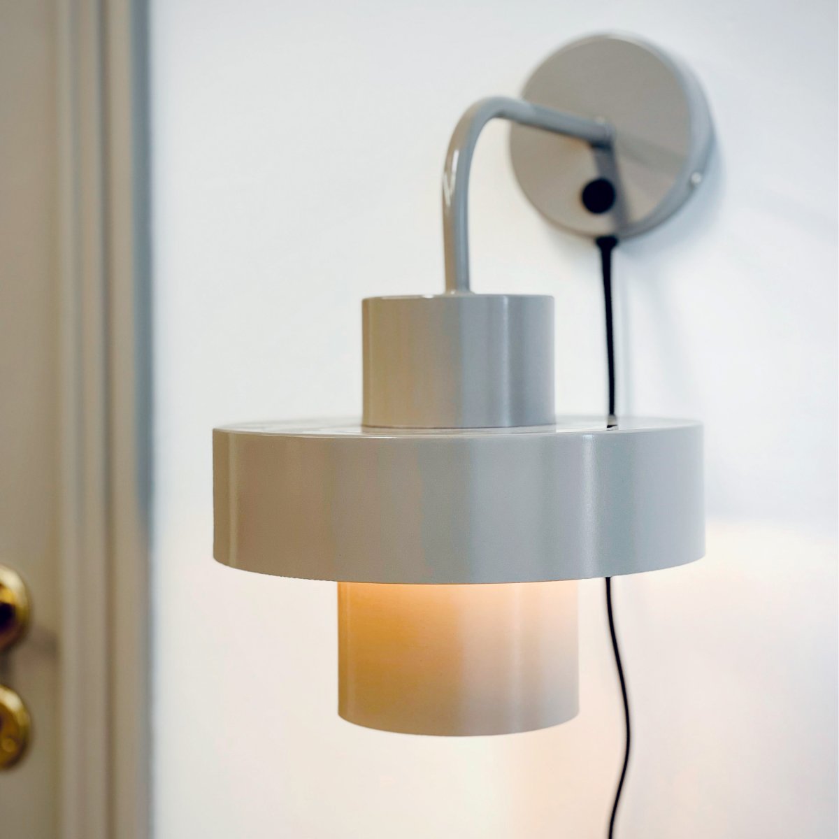 Stoke Ø15 Varmgrå vägglampa från Dyberg Larsen - Den &auml;r tillverkad i metall med en glansig yta som framh&auml;ver lampans cylindriska former och skapar en balanserad kontrast mot andra material som sten, tr&auml; och textilier.
